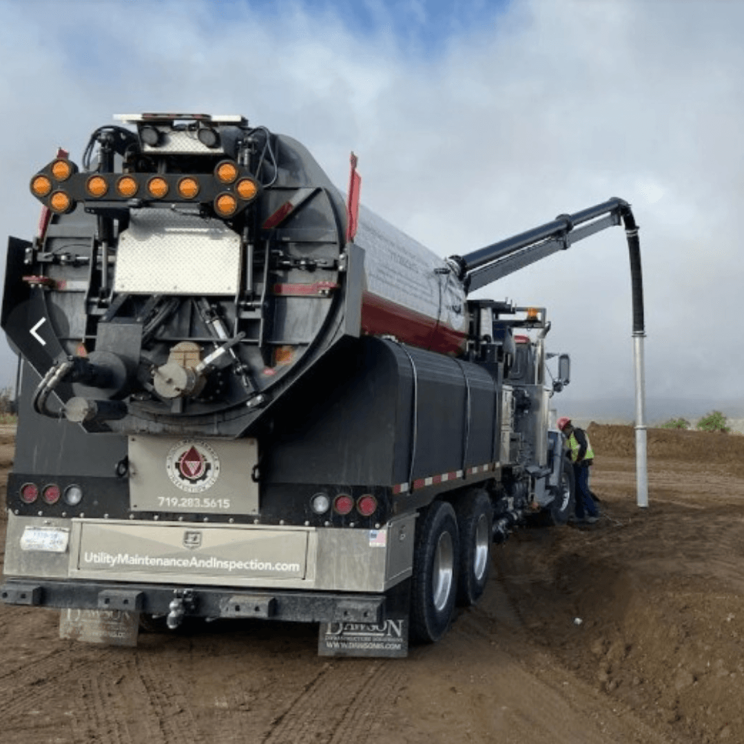 A specialized utility maintenance truck with a large suction tube is set up at a construction site, with two workers in safety gear nearby.
