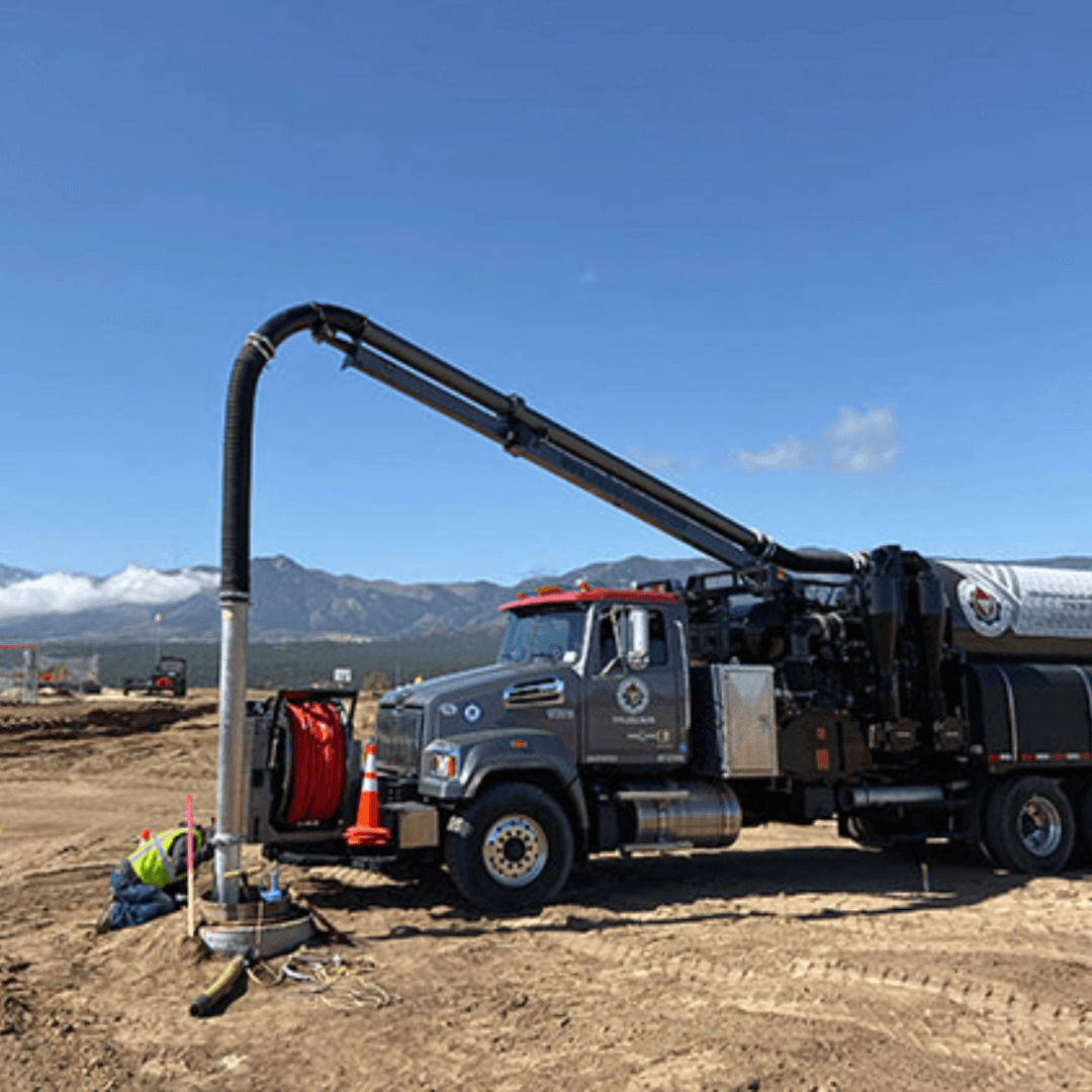 A hydro excavation truck parked on a construction site, featuring a long extended hose and surrounded by equipment on a clear day with mountains in the background.